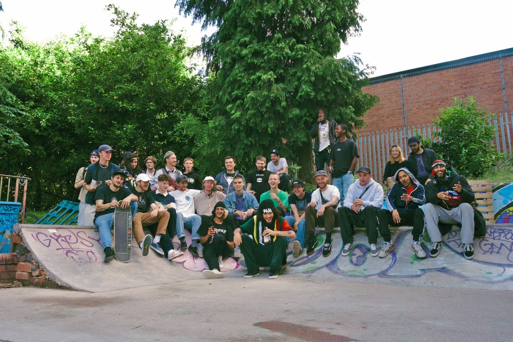 A large group of skateboarders gathered on a DIY skate ramp, smiling and posing for the camera, surrounded by greenery and graffiti.