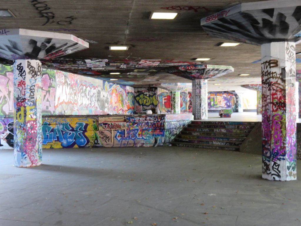 An underground DIY skatepark featuring colorful graffiti on the walls and pillars, with various concrete ramps and ledges for skating.