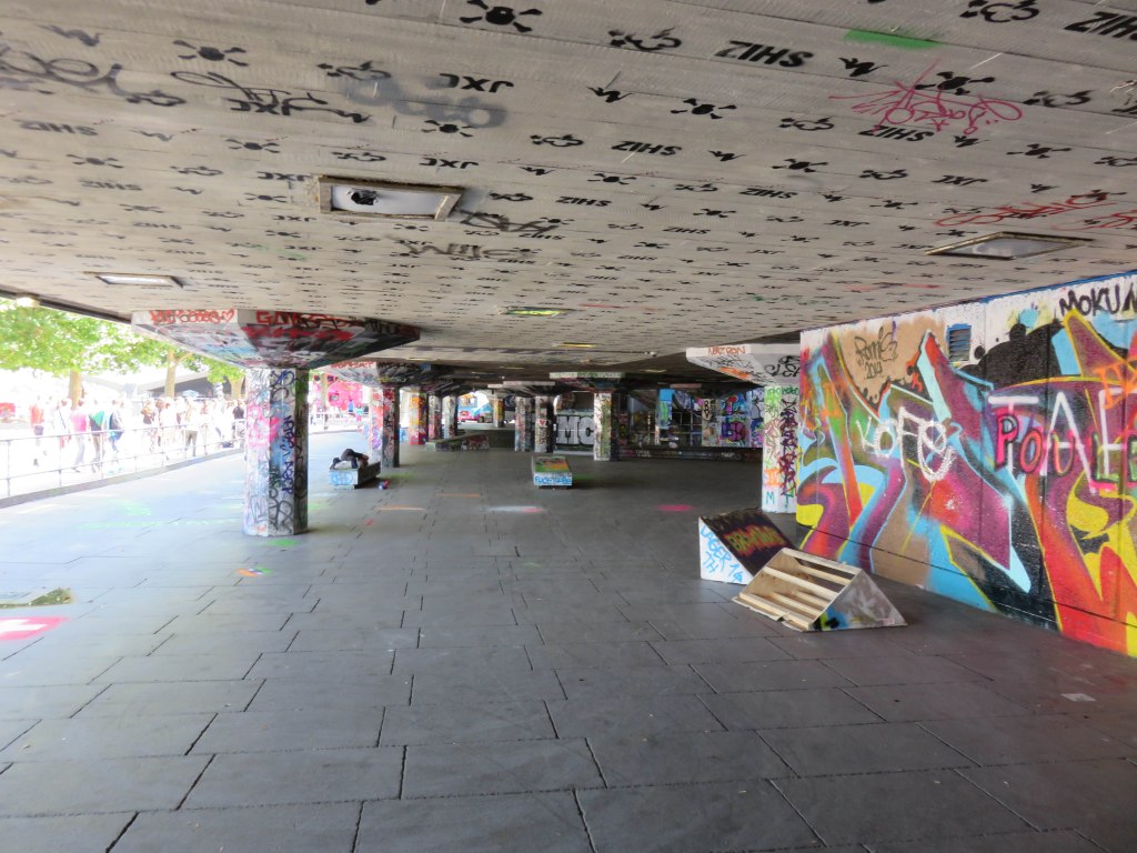 An interior view of a DIY skatepark featuring graffiti-covered walls and columns, with a concrete floor and various makeshift skate obstacles.