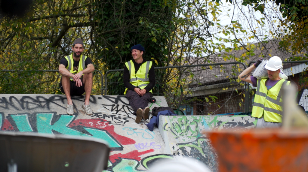 Three individuals in safety vests sitting and working on a DIY skatepark with graffiti-covered surfaces in the background.